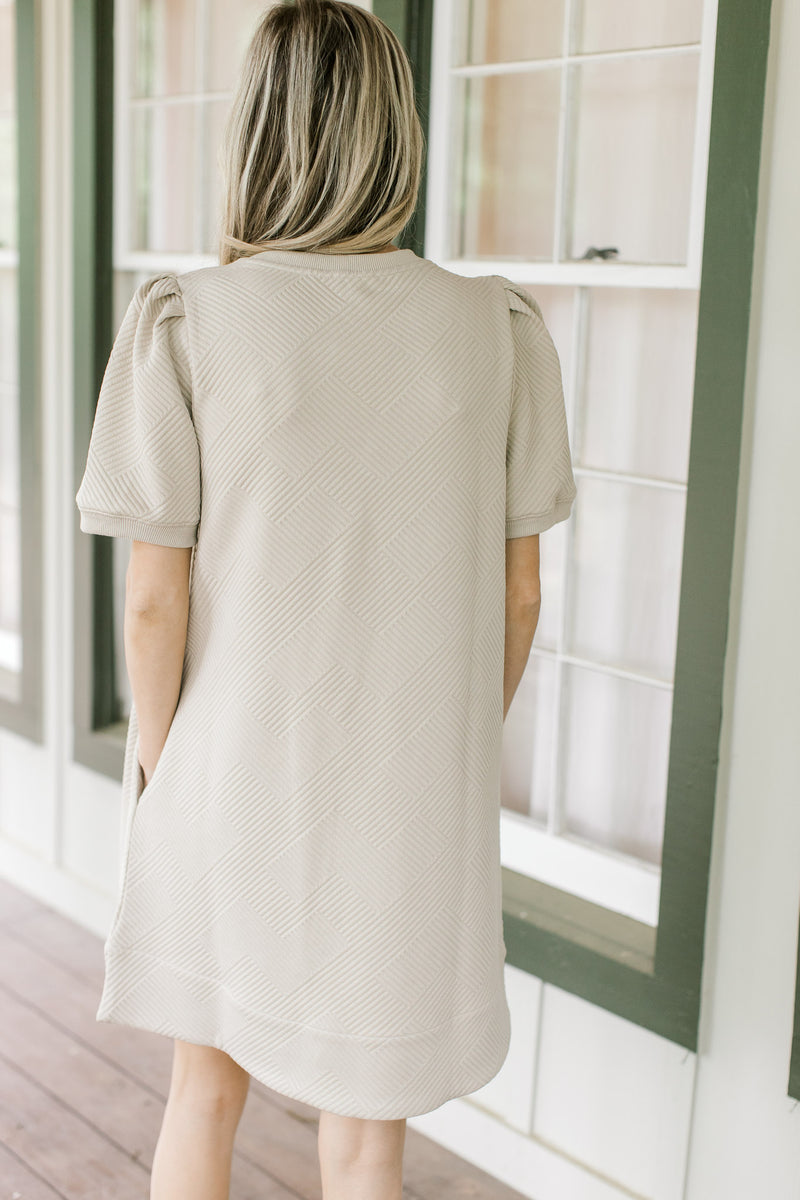 Back view of Model wearing a taupe dress with pleated shoulders and short sleeves.