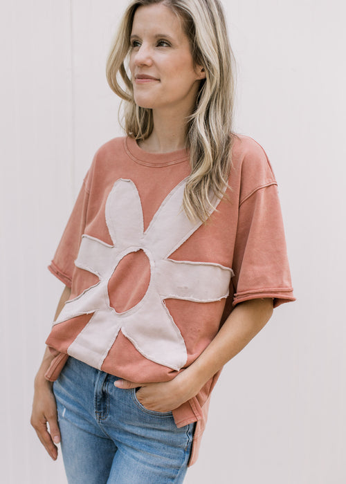 Model wearing a dusty peach top with a cream flower, short sleeves and a split hem. 