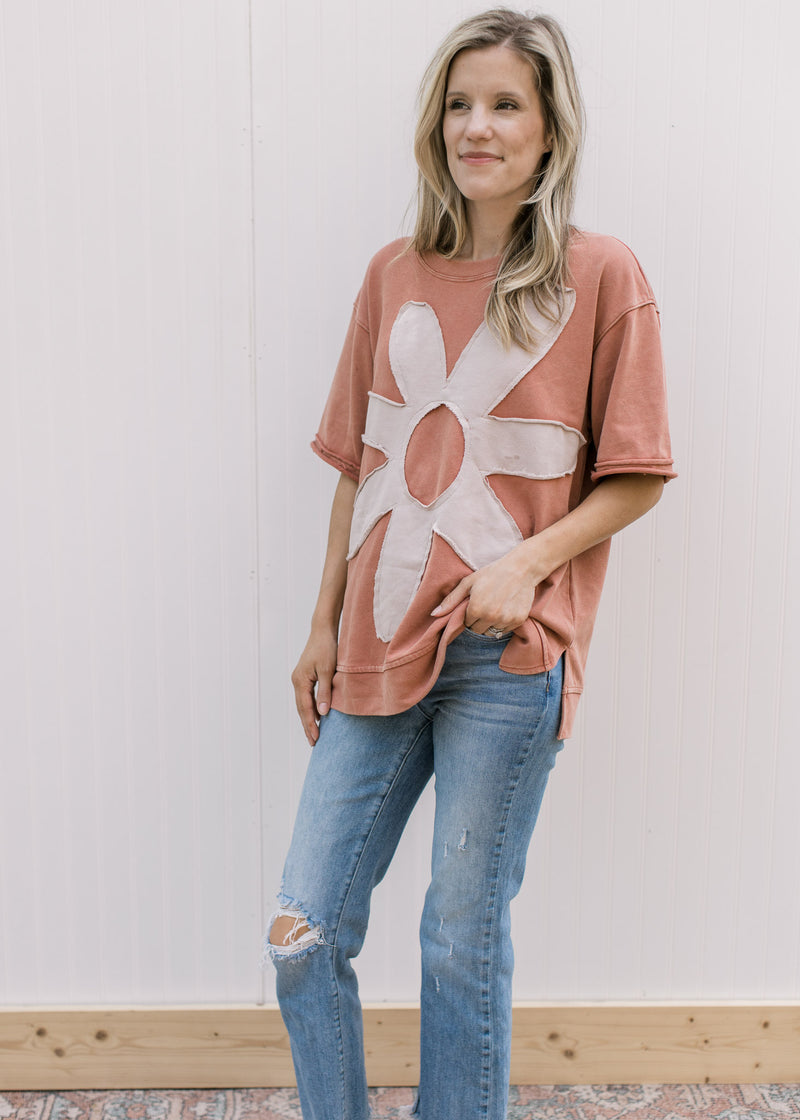 Model wearing a dusty peach top with a cream flower, rolled short sleeves and a split hem.