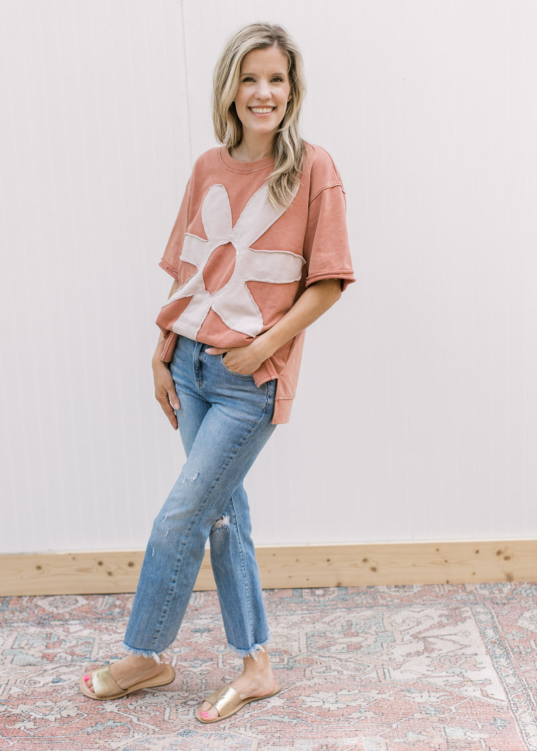 Model wearing jeans and a dusty peach top with a cream flower and short sleeves.