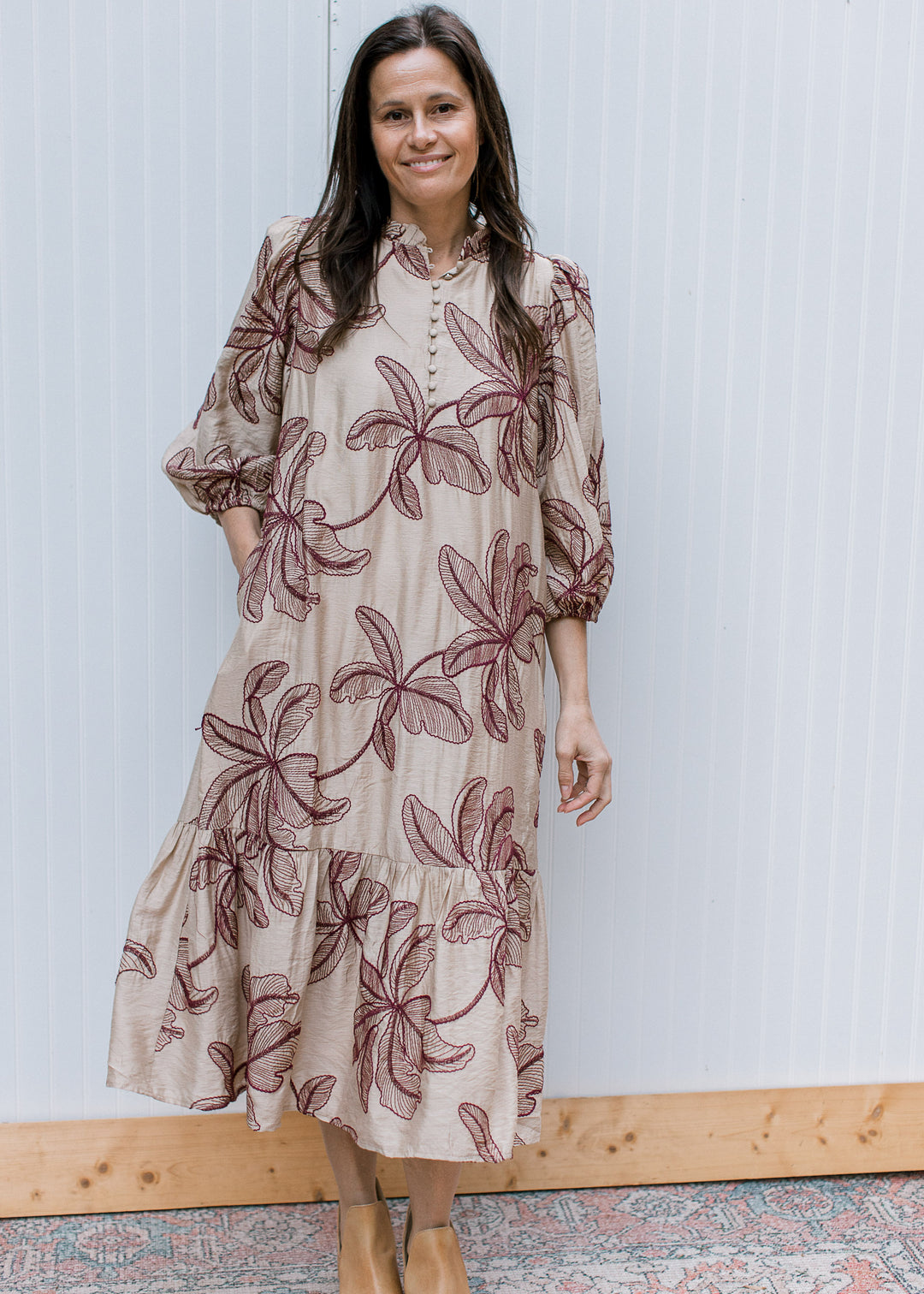 Front view of taupe midi dress with burgundy floral embroidery, button bodice, ruffled hem and hand in side pocket.