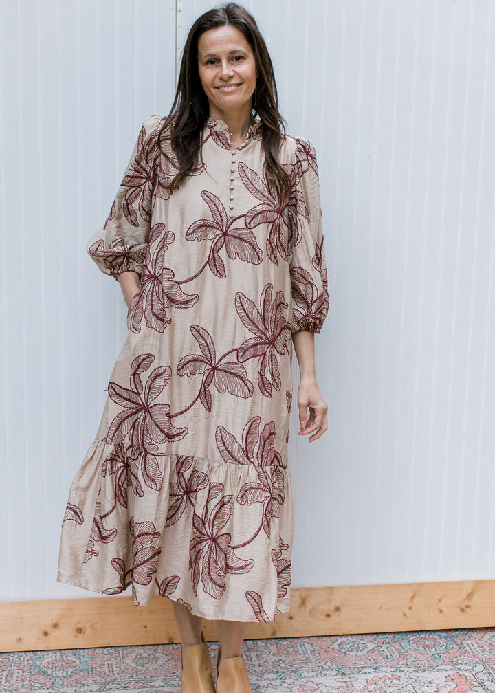 Front view of taupe midi dress with burgundy floral embroidery, button bodice, ruffled hem and hand in side pocket.