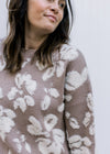 Model wearing a taupe sweater with cream flowers, long sleeves and a slightly distressed hem.