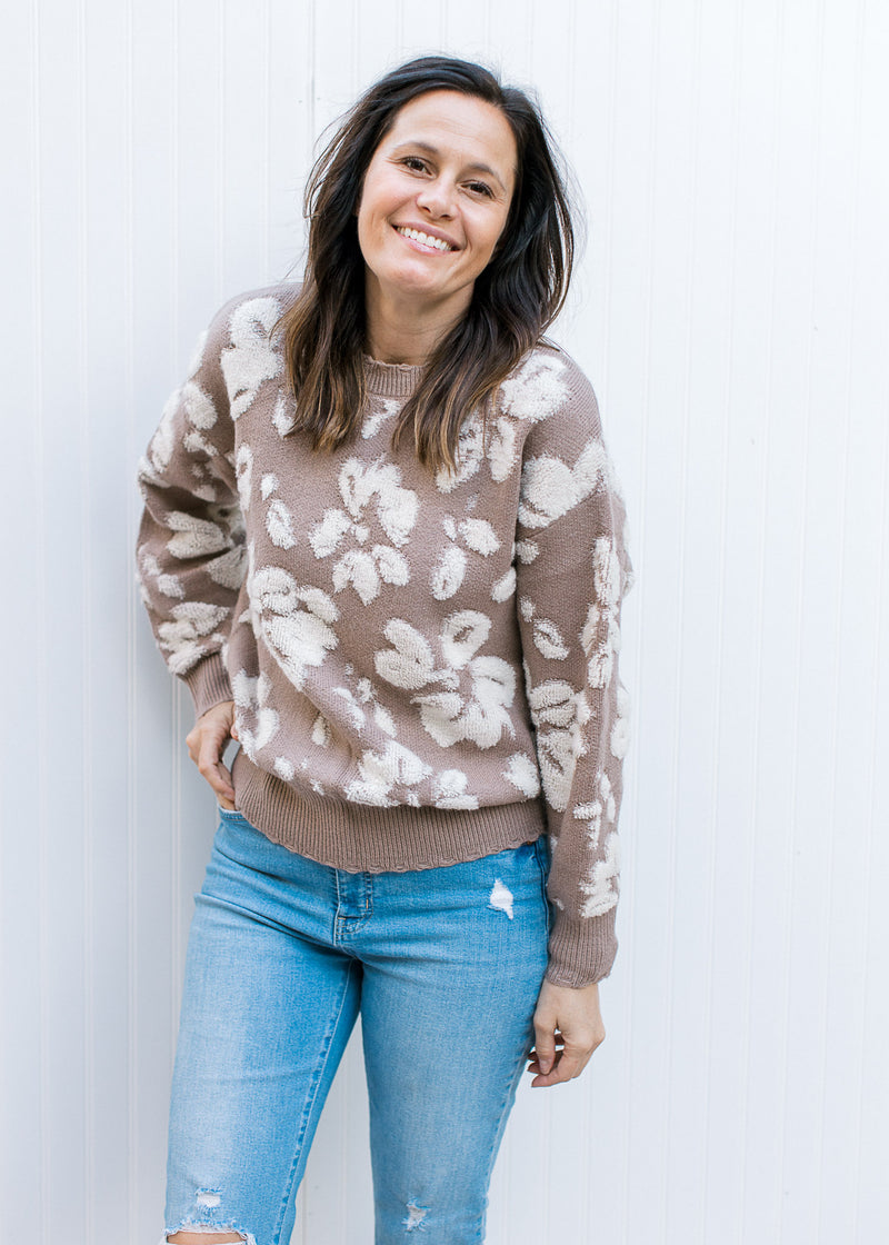 Model wearing jeans and a taupe long sleeve sweater with cream flowers and a distressed hem.