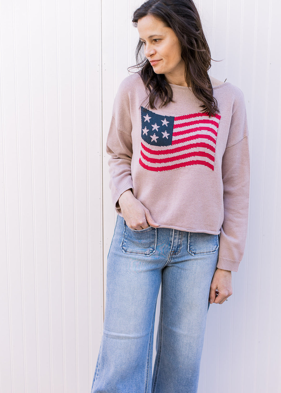 Model wearing a taupe long sleeve sweater with American flag graphic, rolled boat neck and hem.