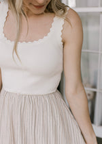 Close up on scalloping on a sleeveless ribbed midi with a cream sweater top and taupe bottom. 