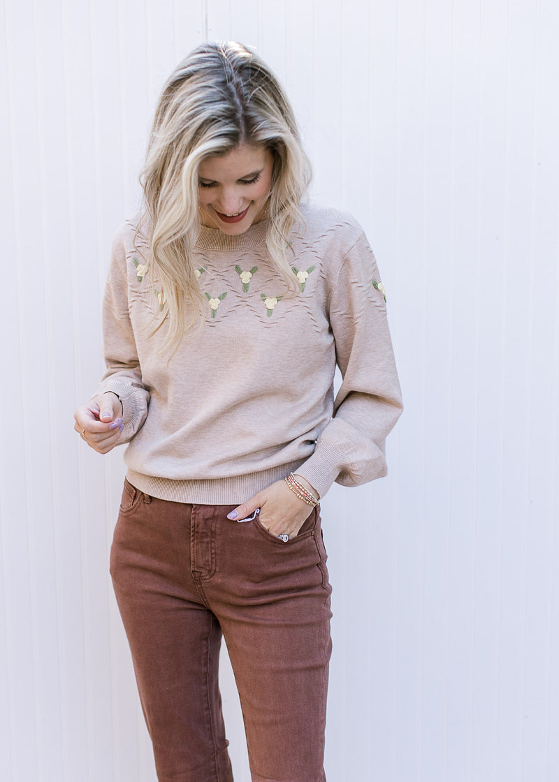 Model wearing brown jeans and a beige sweater with green and yellow flowers and long sleeves.