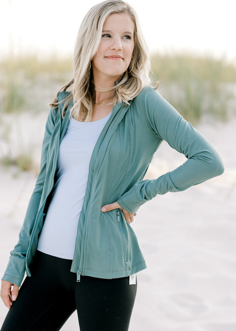 Model wearing a teal, long sleeve, zip up athletic jacket with zipper pockets. 