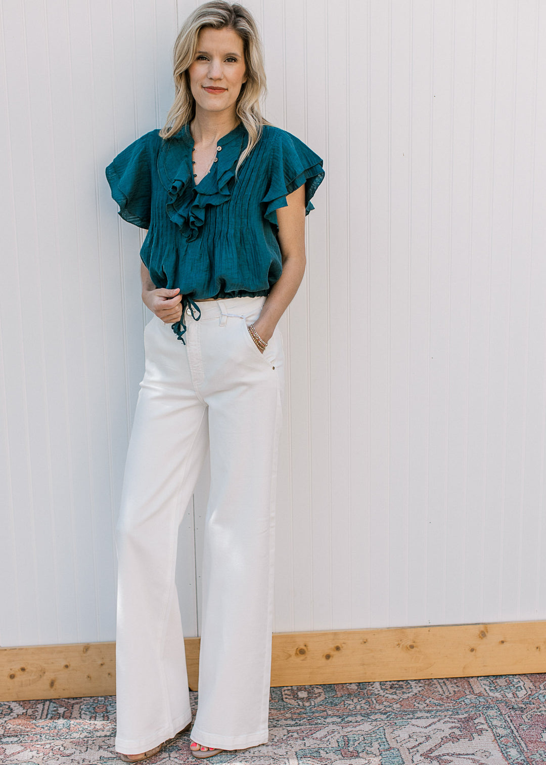 Model wearing white pants and a teal v-neck top with double ruffle and pleated bodice. 