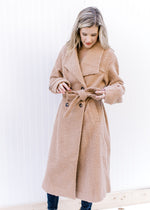 Model wearing a camel, double breasted knee length jacket with a belt and a polyester composure.