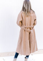 Back view of a Model wearing a camel knee length jacket with a belt and a polyester composure.