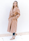 Model wearing booties and a camel, double breasted knee length jacket with belt and long sleeves.