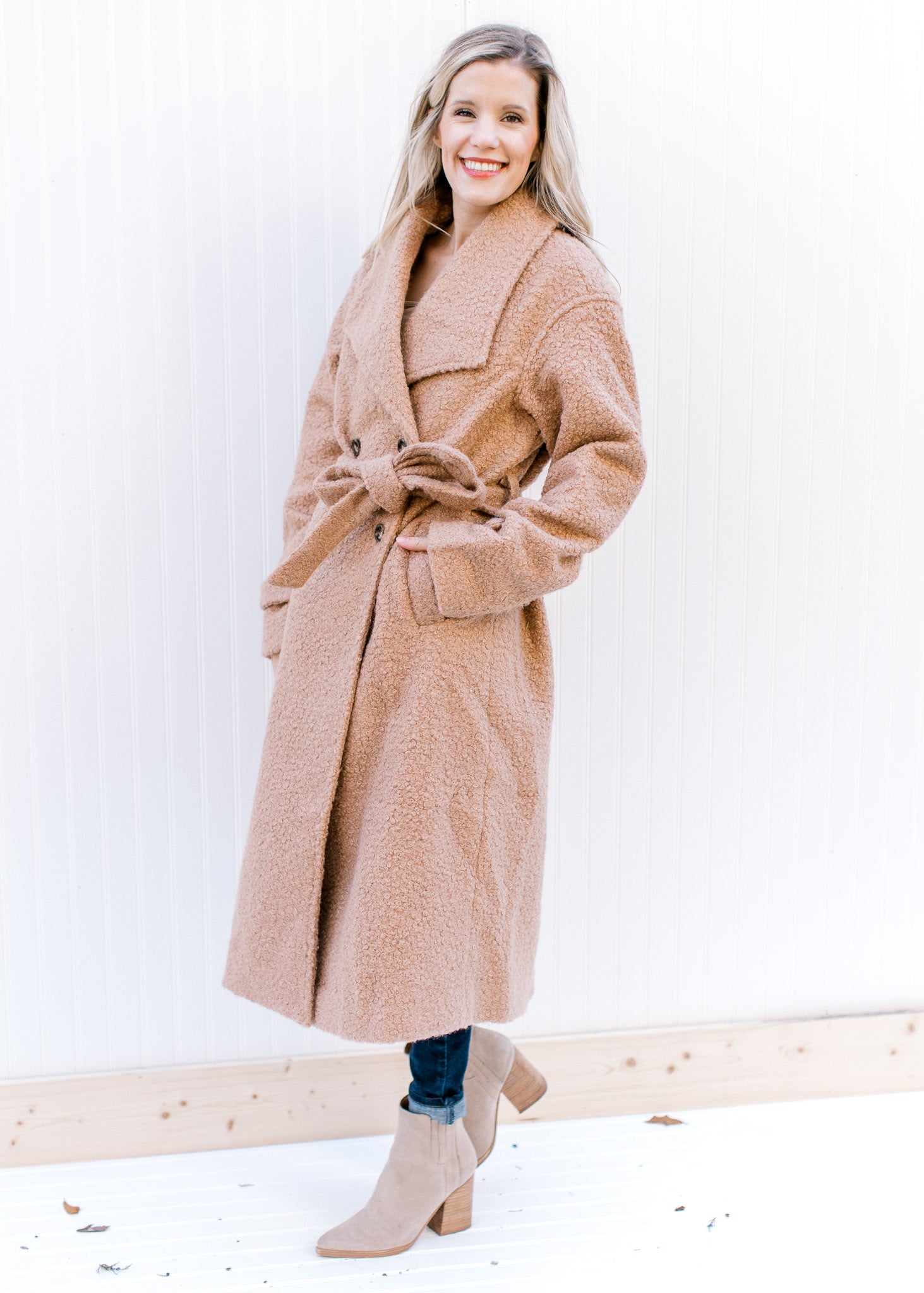 Model wearing booties and a camel, double breasted knee length jacket with belt and long sleeves.