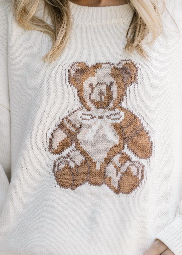 Close up of brown teddy bear on the front of a long sleeve cream sweater with a round neck. 