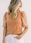 Model wearing a burnt orange top with a cream floral embroidery on scalloped short sleeves. 