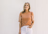 Model wearing white pants with a burnt orange top with a cream floral embroidery. 