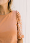 Close up view of cream floral embroidery on scalloped short sleeves of a burnt orange top. 