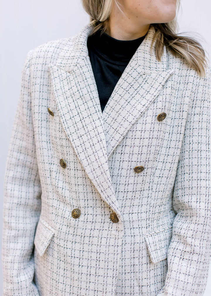 Close up of gold double breasted buttons on a cream plaid blazer with long sleeves and flap pockets.