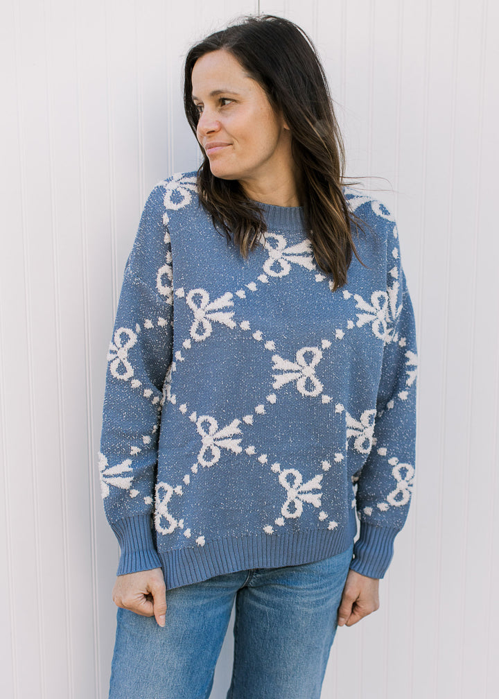 Model wearing a dusty blue sweater with cream ribbons and dots, long sleeves and a round neck. 