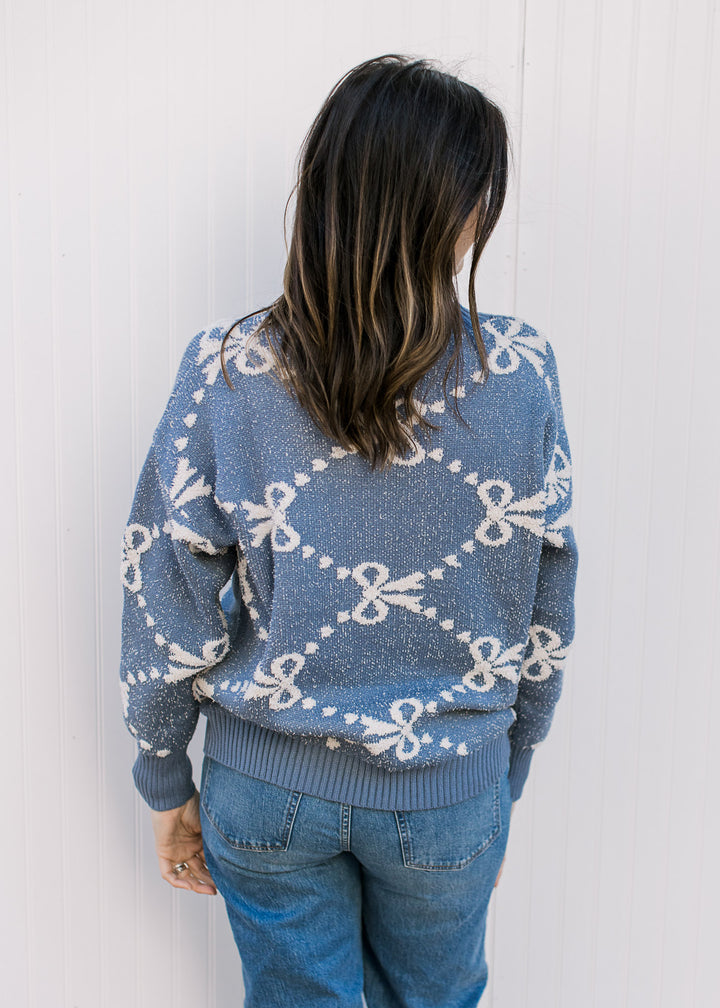 Back view of Model wearing a dusty blue sweater with cream ribbons and dot detail and long sleeves.