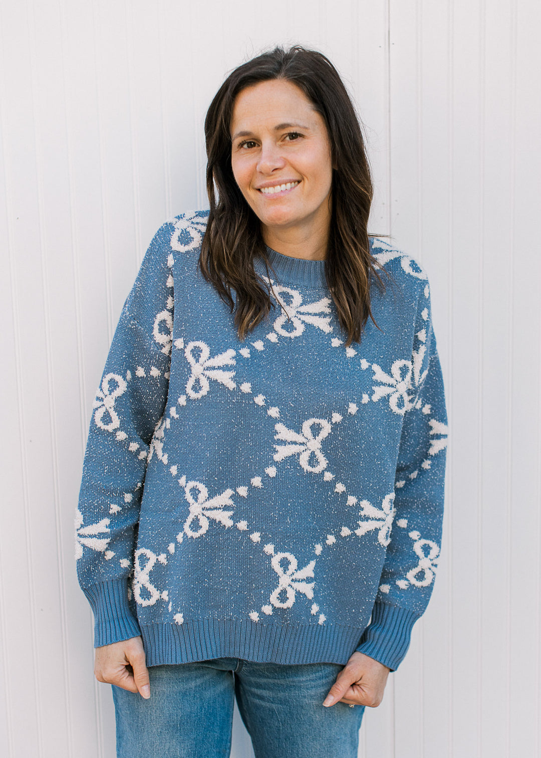 Model wearing jeans and a dusty blue long sleeve sweater with cream ribbons and dot detail.
