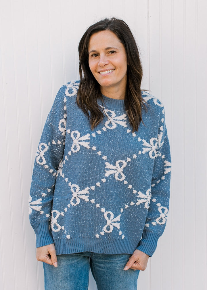 Model wearing jeans and a dusty blue long sleeve sweater with cream ribbons and dot detail.