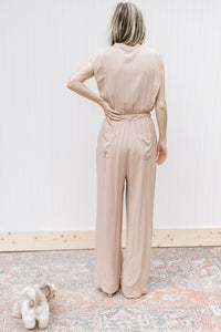 Back view of Model wearing a beige jumpsuit with button bodice, elastic waist and cap sleeves.