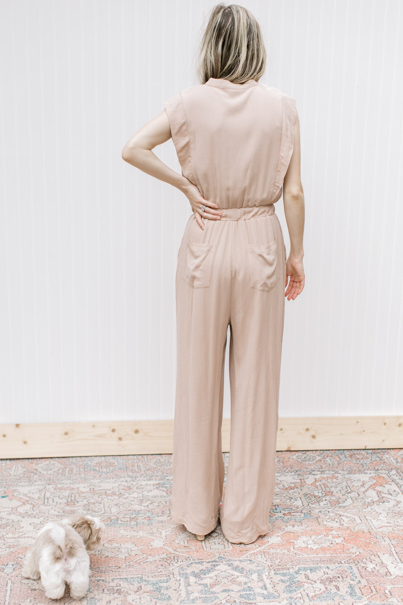 Back view of Model wearing a beige jumpsuit with button bodice, elastic waist and cap sleeves.