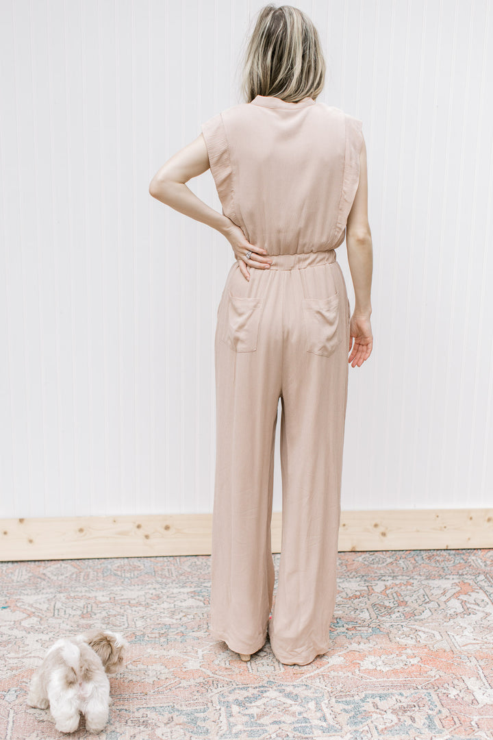 Back view of Model wearing a beige jumpsuit with button bodice, elastic waist and cap sleeves.