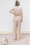 Back view of Model wearing a beige jumpsuit with button bodice, elastic waist and cap sleeves.