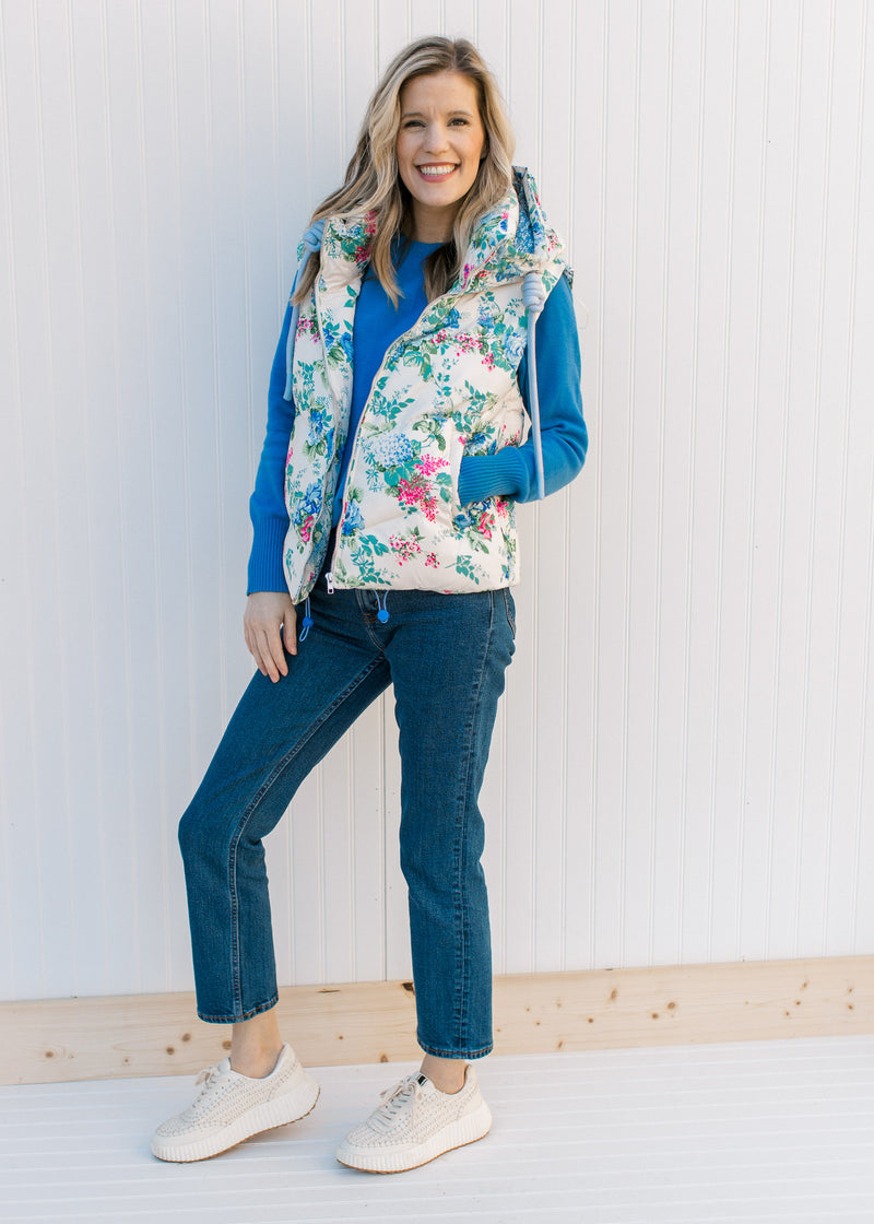 Model wearing jeans, sneakers and a white puffer vest with blue, pink and green floral design. 
