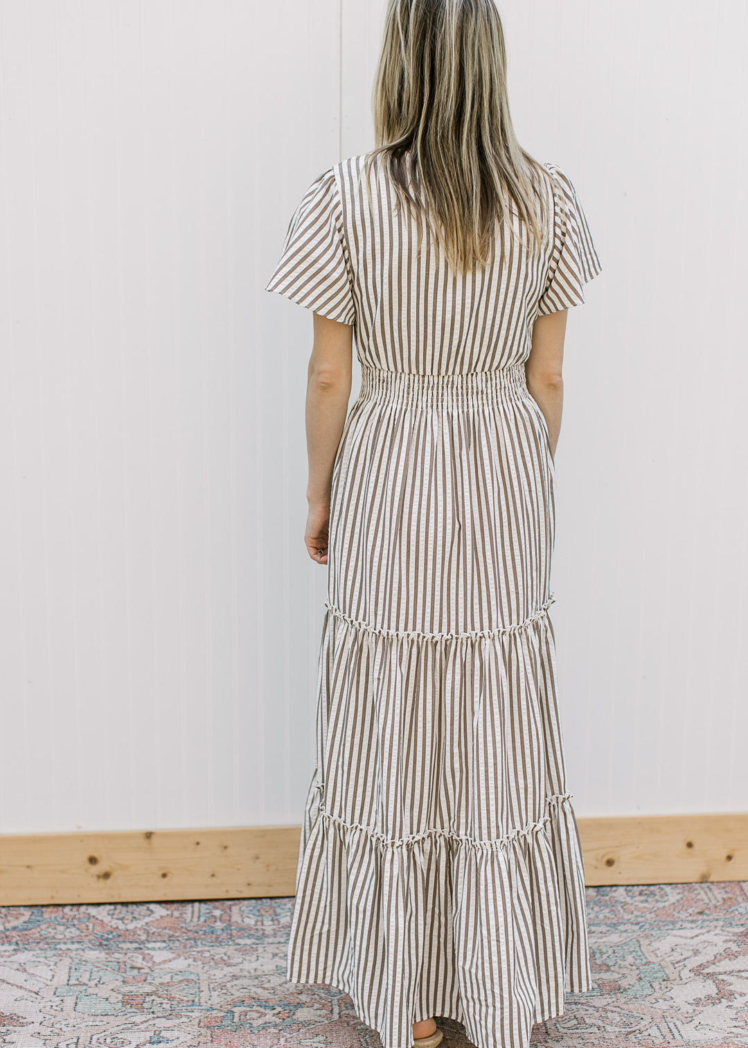 Back view of Model wearing a cream maxi with brown stripes, v-neck, short sleeves and elastic waist.
