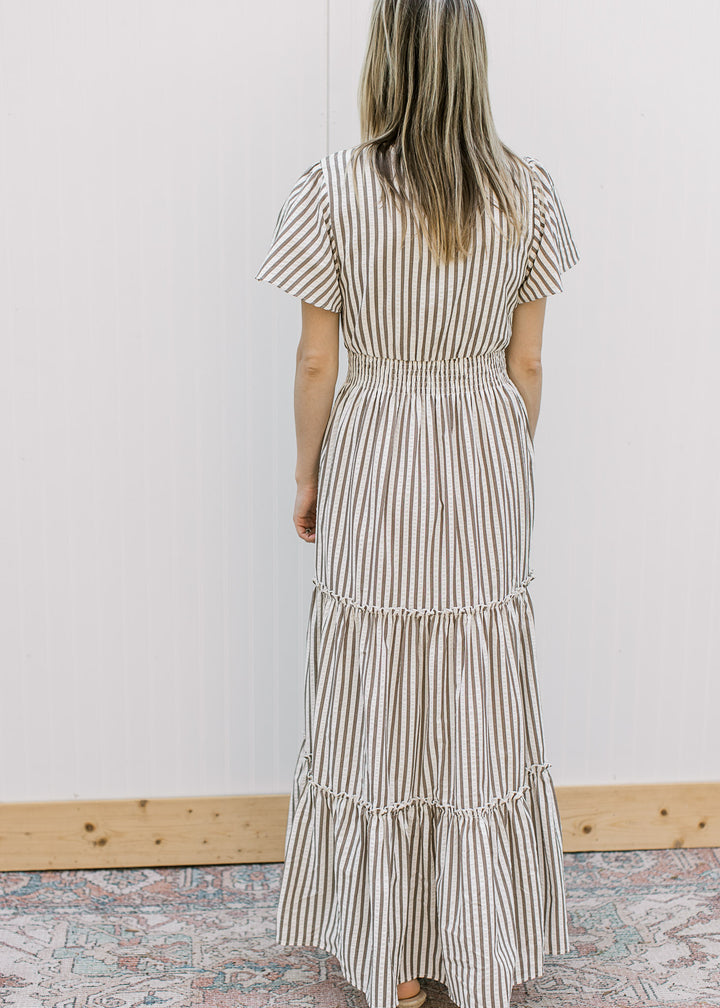 Back view of Model wearing a cream maxi with brown stripes, v-neck, short sleeves and elastic waist.
