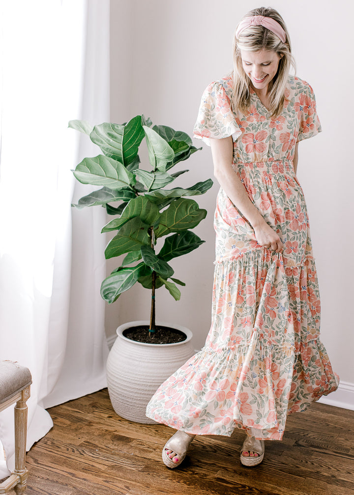 Model wearing a beige maxi with pink, floral, flutter short sleeves, v-neck and tiered skirt. 