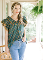 Model wearing a teal top with gold flowers, gold scalloped lace and a flap collar. 