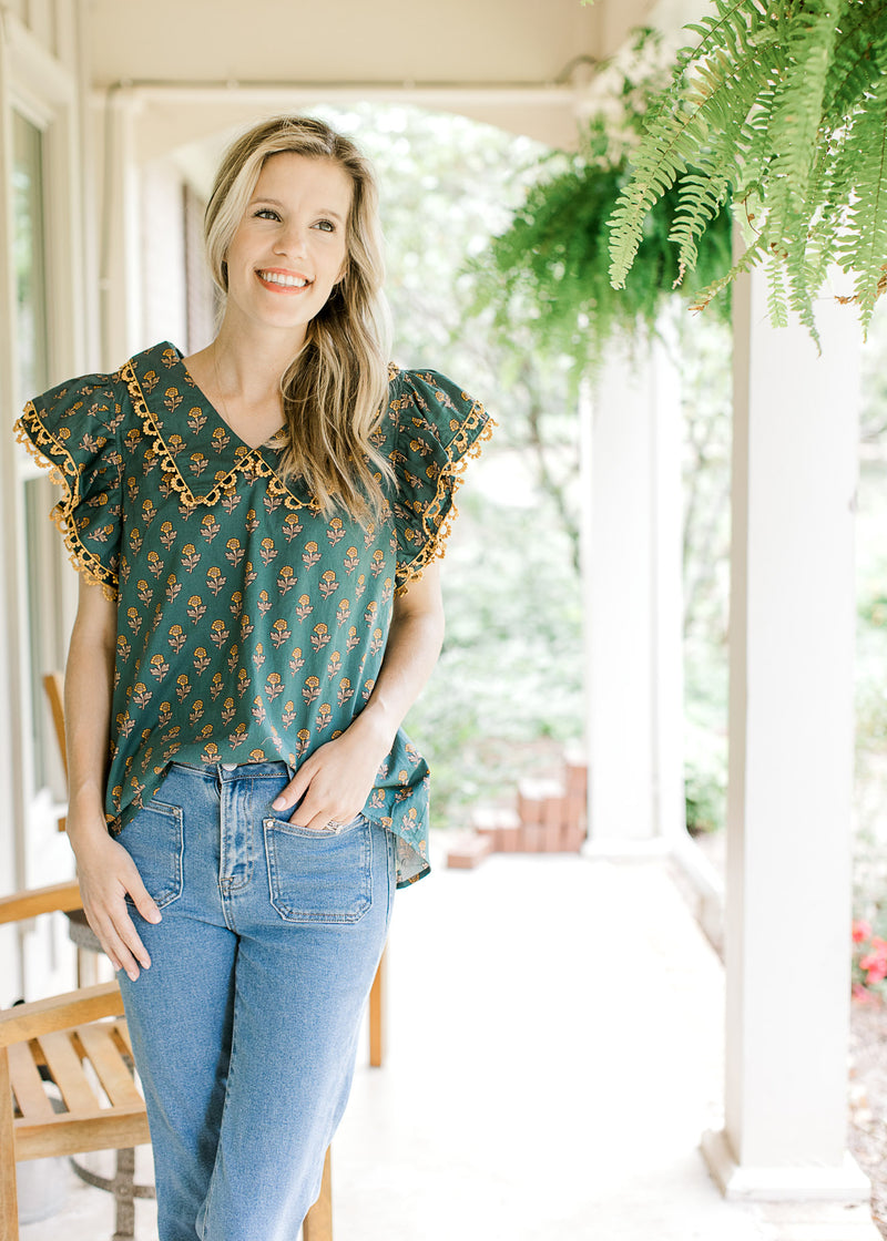 Model wearing a teal top with gold flowers, layered short sleeves and a flap collar. 