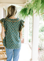 Back view of Model wearing a teal top with gold flowers, gold scalloped lace and a flap collar. 