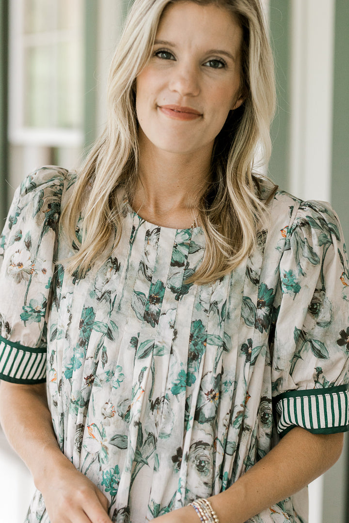 Model wearing a grayish tan dress with green floral, round neck with pleating and short sleeves. 