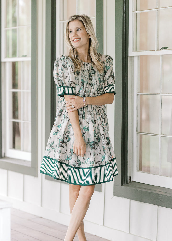 Model wearing a grayish tan dress with green floral, round neck  and bubble short sleeves. 