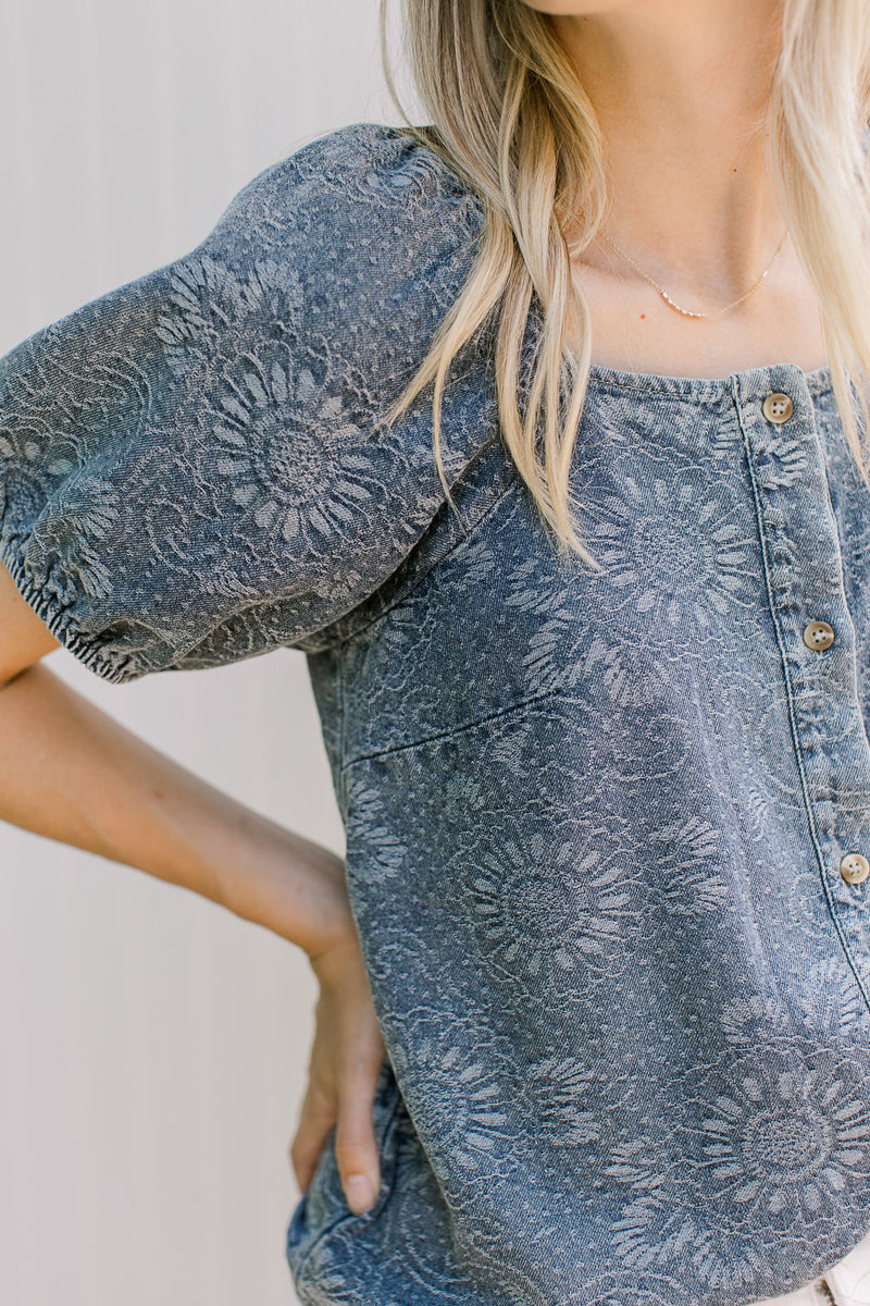 Close up of floral pattern on a blue button up top with a square neck and short sleeves. 