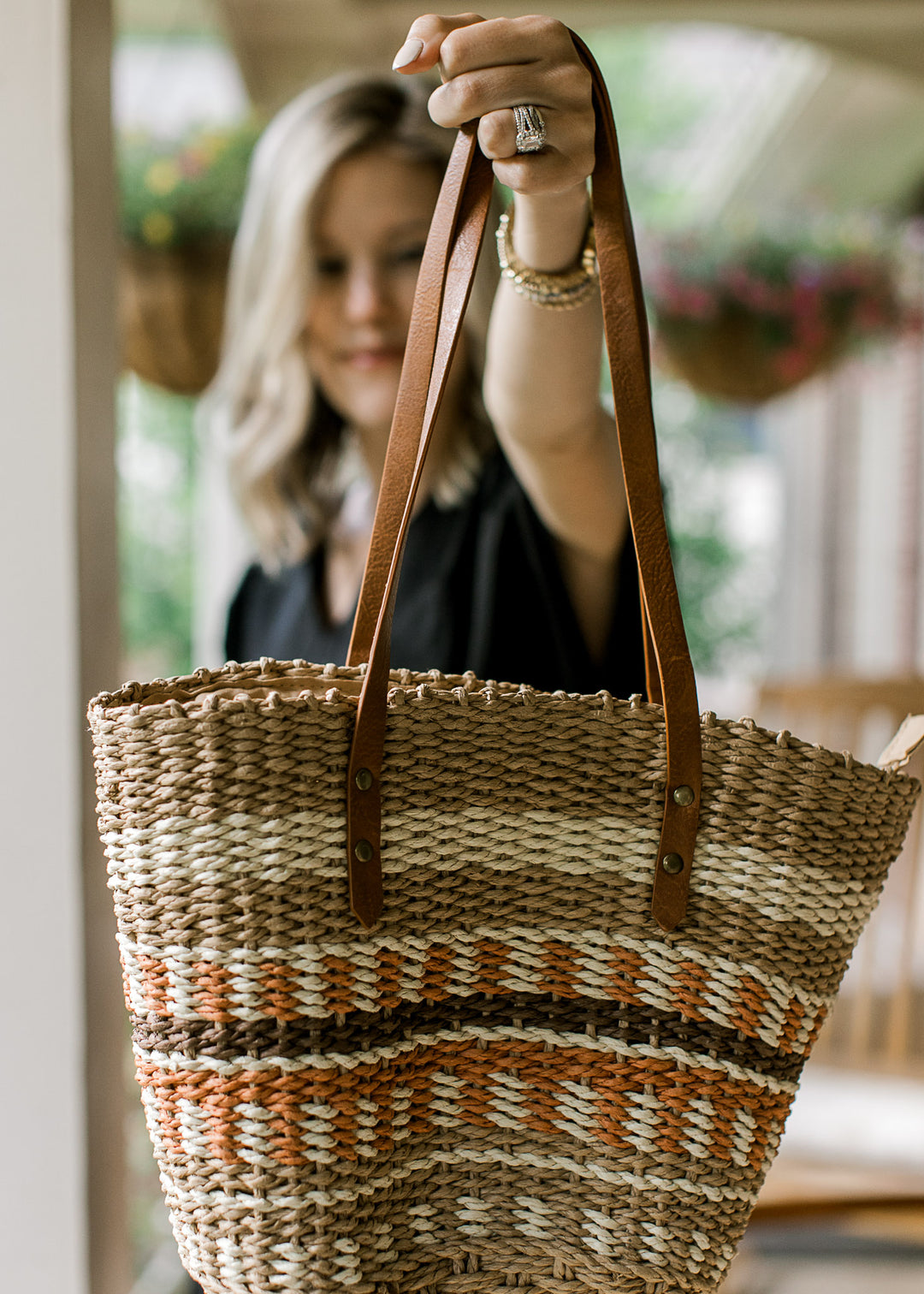 Model holding a woven straw tote with taupe, cream, brown, and rust pattern and faux leather straps.