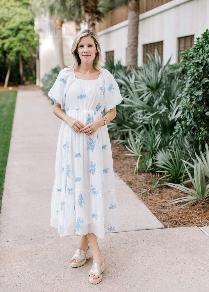 Model wearing wedges and a white midi with pale blue flowers, square neck and short bubble sleeves.