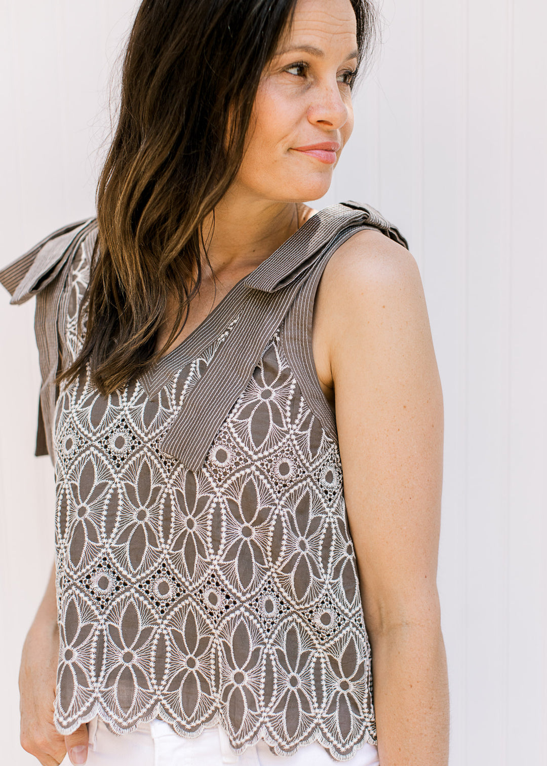 Model wearing a mocha v-neck top with cream floral embroidery, faux tie straps and a scalloped hem.