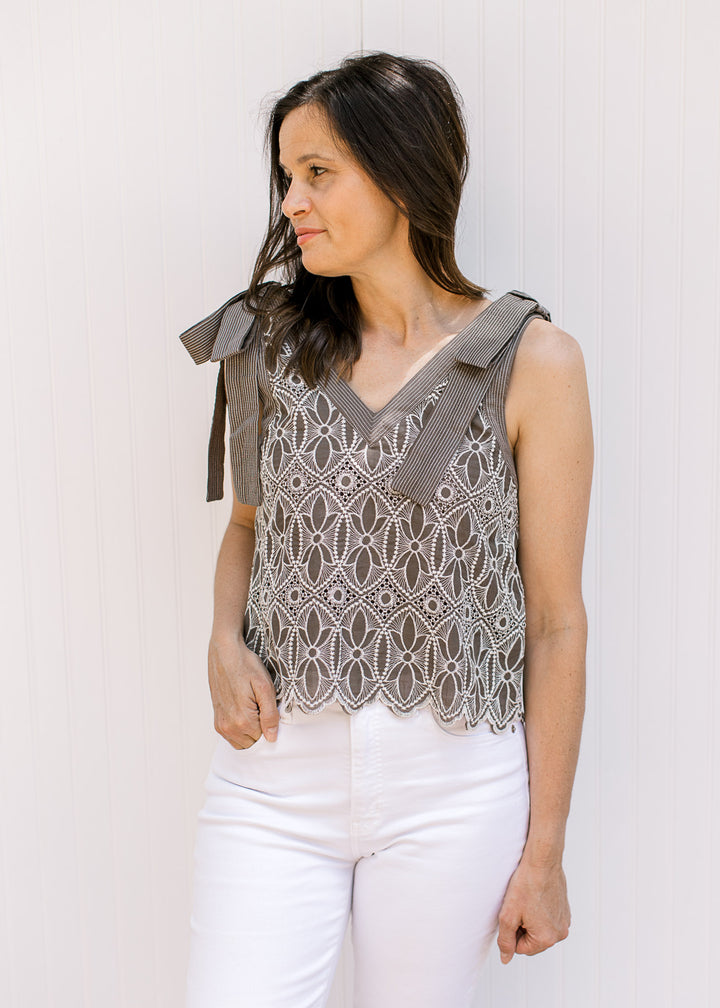 Model wearing white pants and a mocha top with cream floral embroidery and faux tie straps. 