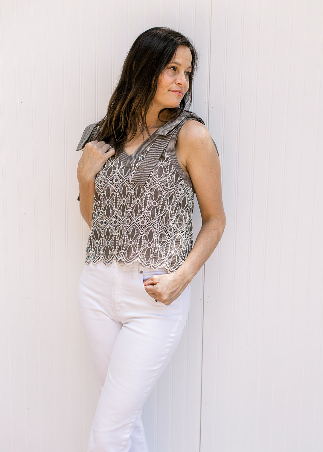 Model wearing white pants and a mocha top with cream floral embroidery and faux tie straps. 