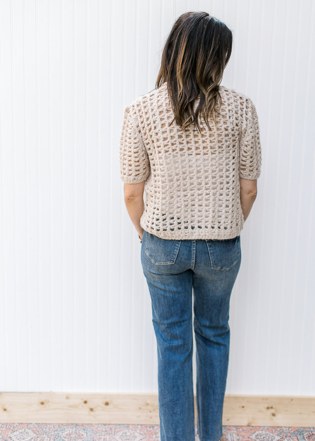 Back view of Model wearing a oatmeal colored, open crochet cardigan with short sleeves.