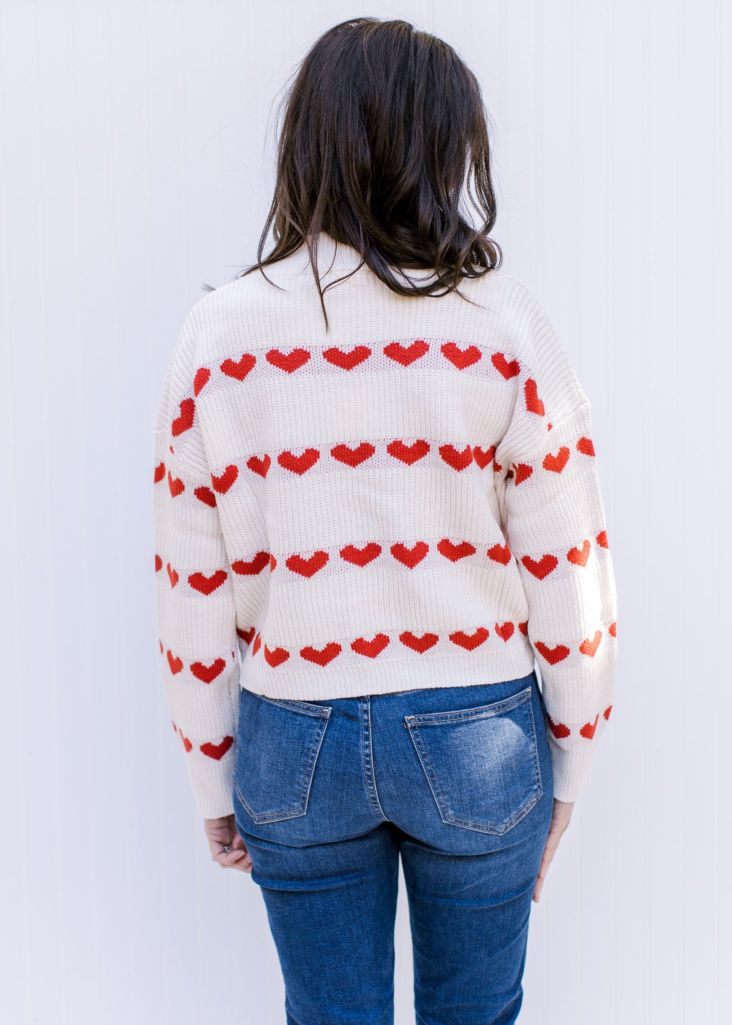 Back view of Model wearing a cream sweater with a heart pattern, mock neck and long sleeves. 