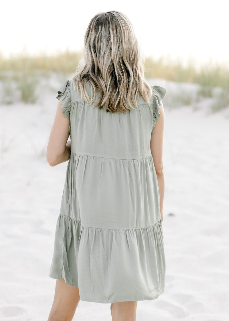 Back view of Model wearing a tiered, above the knee, olive dress with flutter cap sleeves. 