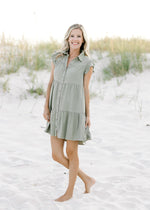 Model wearing an above the knee, rayon, olive dress with flutter cap sleeves and button closure.