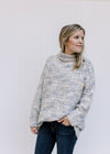 Model wearing a gray, cream and mustard tweed sweater with a turtleneck, long sleeves and pockets. 